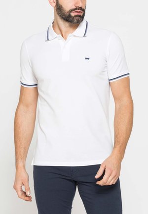 Uomo che indossa una polo bianca con rifiniture blu navy su maniche e colletto, abbinata a pantaloni blu scuro, in piedi davanti a uno sfondo semplice.