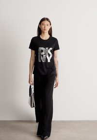 Mulher com cabelos longos e escuros, vestindo uma t-shirt preta da DKNY e calças largas pretas, carregando uma mala prateada, a caminhar no interior sobre um chão cinzento.