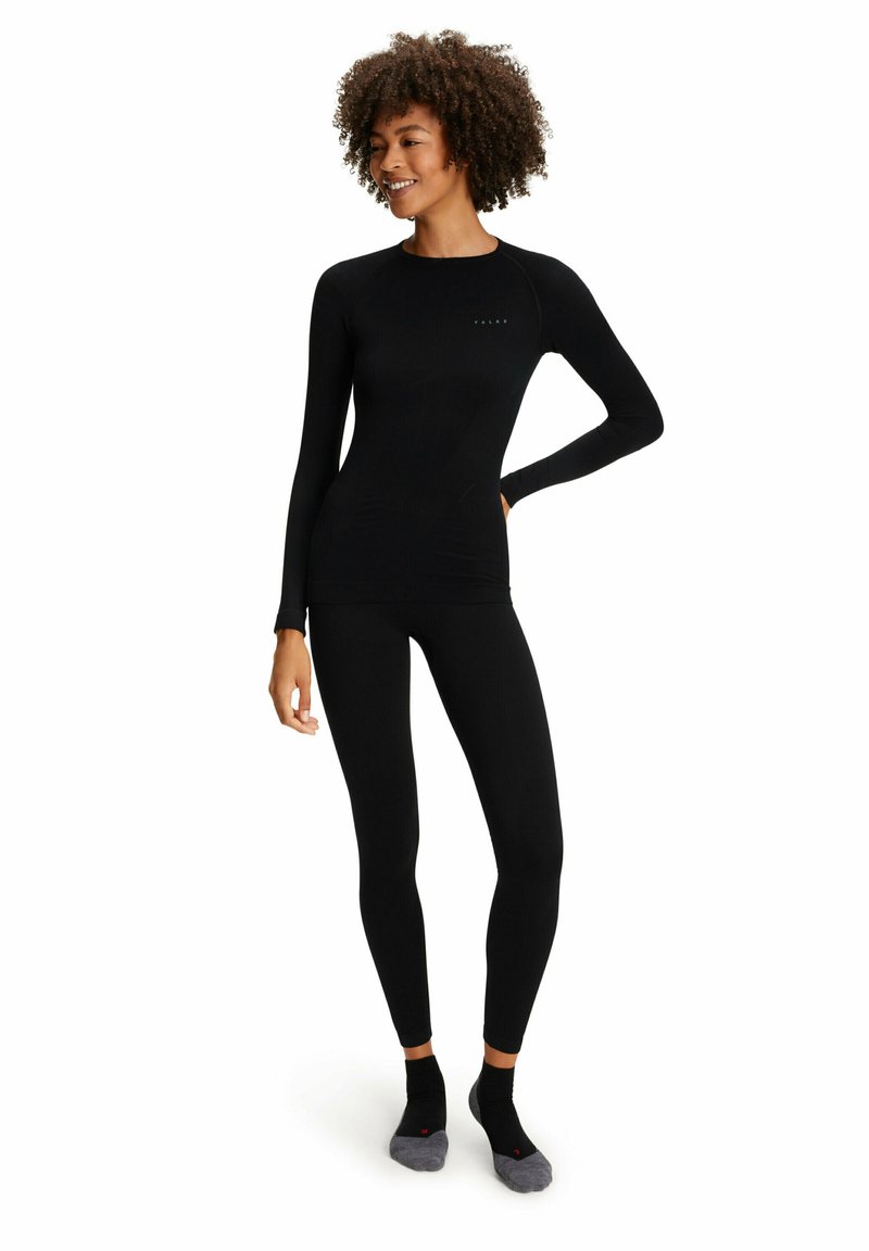 Schwarz langärmliges Oberteil und enganliegende Leggings in geripptem Material, mit einem kleinen Logo auf der Brust und weichen, knöchellangen Socken.