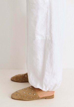 Pies con zapatos marrones tipo slip-on con tachuelas metálicas redondas, combinados con pantalones blancos holgados con puños elásticos.