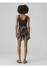 Žena s kudrnatými vlasy, oblečená v námořnické bezramínkové topu a vysokých námořnických šortkách s mnohobarevným vzorem listů, stojící s otočením zády.