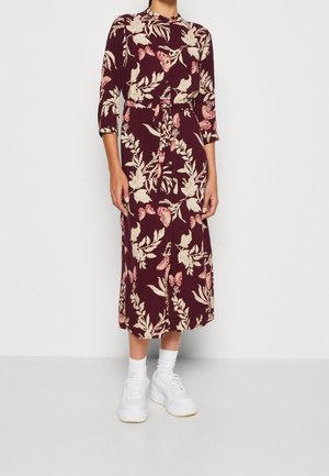 Nainen pukeutuneena burgundinpunaisiin midi-mittaisiin mekkoon, jossa on beige kukka- ja perhosteema, kolmen neljänneksen hihat, vyötärönauha, valkoiset sukat ja valkoiset tennarit.