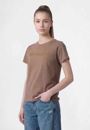 Junge Frau mit braunem hohem Pferdeschwanz trägt braunes Champion-T-Shirt und blaue Jeans mit kleinem Riss, steht mit einer Hand in der Tasche.