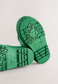Scarpe in gomma verdi con suole testurizzate caratterizzate da un motivo giocoso a schizzi e una faccina sorridente. Progettate per durabilità e aderenza.