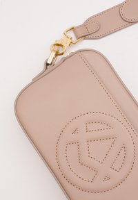 Custodia per telefono in pelle rosa chiaro con logo impresso, hardware dorato e una tracolla staccabile. Texture liscia con dettagli cuciti.