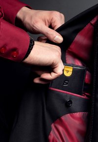 Black blazer with red textured lining; a hand is placing a black wallet with a yellow card into an interior pocket. Red shirt with buttons visible.
