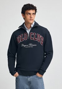 Marinblå fleece-tröja med hög krage, halvvajer och "POLO CLUB" broderat i vinröd. Bärs över en ljusblå skjorta.
