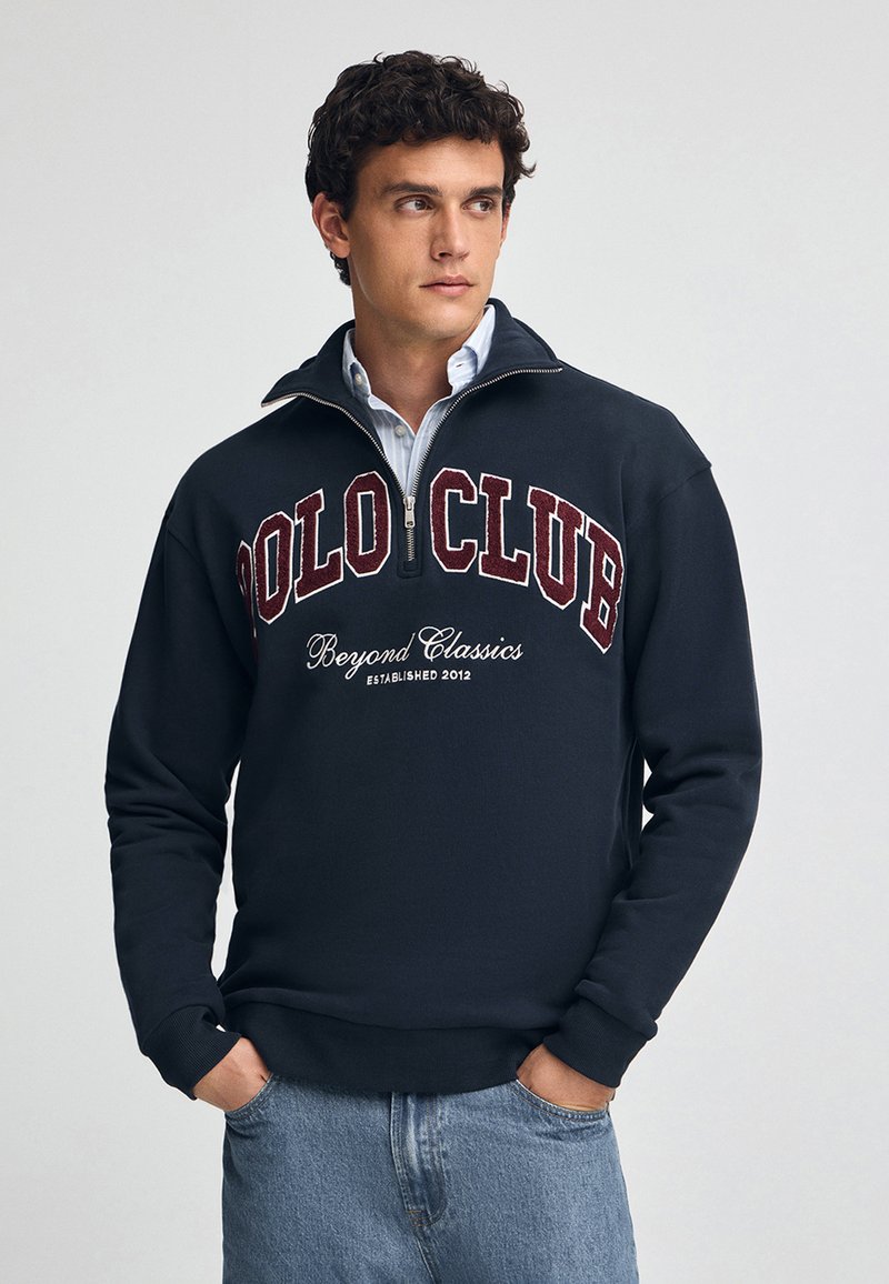 Marinblå fleece-tröja med hög krage, halvvajer och "POLO CLUB" broderat i vinröd. Bärs över en ljusblå skjorta.