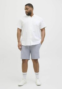 Chemise blanche à manches courtes avec un motif texturé et une poche, associée à un short bleu clair. Baskets blanches, chaussettes blanches avec des lettres noires.