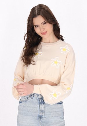 Tânără cu păr lung și castaniu zâmbește, purtând un pulover cropped bej deschis cu modele de margarete albe și galbene și blugi albaștri cu talie înaltă.