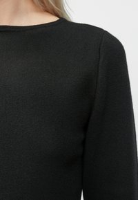 Lähi kuva henkilöstä, joka on pukeutunut yksinkertaiseen mustaan neuleeseen, näyttää ylärintakehän, olkapään ja kaula-aukon.