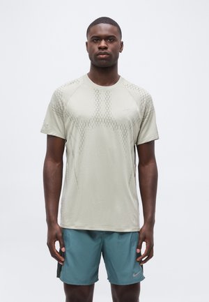 Jeune homme portant un t-shirt de sport gris clair à manches courtes et un short de sport bleu sarcelle, debout, les bras détendus le long du corps, sur un fond uni.