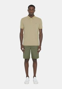 Polo shirt khaki con maniche corte, pantaloni shorts verdi e sneakers bianche. Presenta un discreto logo sul petto. Tessuto liscio, vestibilità su misura.