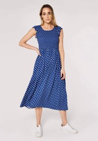 Blue polka dot dress with ruffled cap sleeves, fitted bodice, and a flowing midi skirt. Worn with white sneakers for casual contrast.