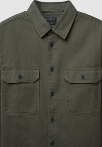 Camicia verde oliva con colletto a polo, due tasche sul petto, chiusura a bottoni e tessuto testurizzato. Presenta bottoni neri e cuciture dritte.