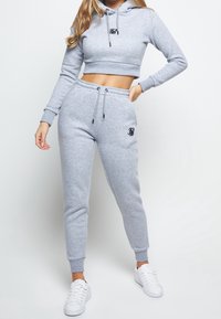 Sudadera corta en gris claro con un logo en la parte frontal, combinada con pantalones jogger a juego que cuentan con una cinturilla elástica y puños. Zapatillas blancas completan el look.