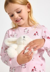 Biały pluszowy królik trzymany przez dziecko w różowej bluzce z długim rękawem ozdobionej postaciami z kreskówek i sercami, o gładkiej fakturze i miękkim materiale.