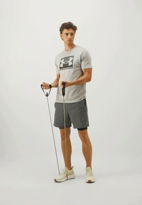 Sivasti športna majica z logotipom, temni šortsi, drži odporne trakove z črnimi ročaji, obut v svetle teniske, stoji na enobarvni podlagi.