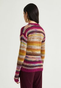 Donna con capelli scuri che indossa orecchini statement dorati e un maglione a righe soffice nei toni del rosa, giallo, marrone e blu, vista da dietro.