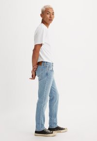 Un tânăr cu părul scurt blond, îmbrăcat cu un tricou alb, blugi deschiși la culoare și pantofi sport negri, stând cu mâinile la spate.