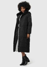 Manteau long rembourré noir avec une capuche doublée de fourrure, des poches latérales et une fermeture éclair. Le modèle porte une robe grise et des bottines noires.