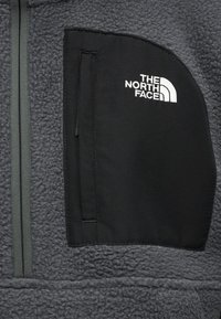 Donkergrijze fleecejas met verticale rits en zwarte borstzak met wit The North Face-logo.