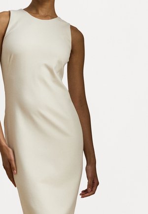 Kobieta w obcisłej, bezrękawnikowej sukni w kolorze kremowym, stojąca na tle jednolitego, jasnego tła.