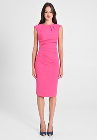 Figurbetontes, ärmelloses Kleid bis zum Knie aus leuchtend pinkfarbener Stoff mit Raffung. Kombiniert mit dunkelblauen Peeptoe-Absätzen.