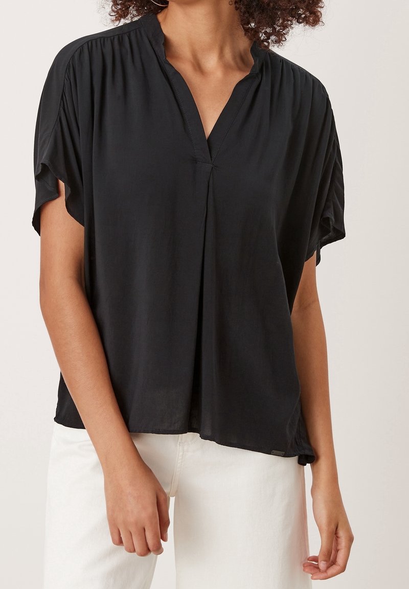 Femme portant un chemisier noir ample à manches courtes avec un col en V et un pantalon blanc, se tenant devant un fond clair uni.