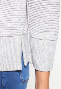 Grauer, gerippter Pullover mit einem High-Low-Saum, strukturiertem Stoff und seitlichen Schlitzen, kombiniert mit blauen Denim-Jeans.