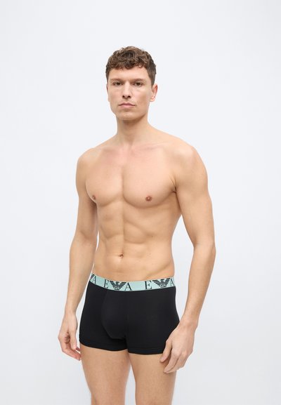 Άνδρας χωρίς πουκάμισο, όρθιος και στραμμένος προς τα εμπρός, φοράει μαύρα κοντά εσώρουχα τύπου μποξεράκι με ανοιχτό μπλε ελαστική μέση που εμφανίζει επαναλαμβανόμενο μοτίβο λογότυπου.