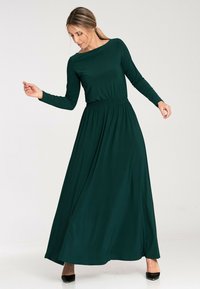Długa, ciemnozielona sukienka maxi z długim rękawem o luźnym kroju, z elastycznym pasem w talii i gładką teksturą tkaniny. Charakteryzuje się dekoltem w łódkę.