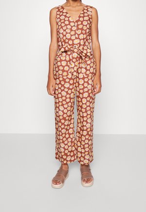 Combinaison marron à motif floral avec des marguerites jaunes, encolure en V, haut sans manches, pantalon à jambes larges, associée à des sandales marron à semelle épaisse.