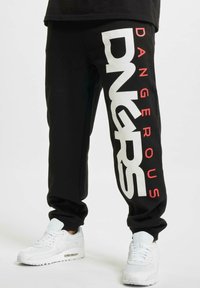Pantaloni sportivi neri con bold lettering bianco e rosso che dice "DANGEROUS" sui lati. Vita e polsini in elastico. Abbinati a sneakers bianche.