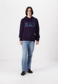 Felpa viola con logo "GAP" blu, tasca frontale, abbinata a jeans azzurri chiari e sneaker nere con strisce bianche, esposta su uno sfondo semplice.
