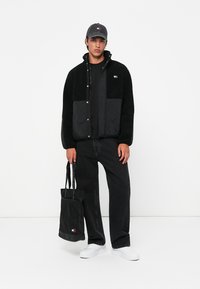 Zwarte fleecejack met gestikte panelen, zwart t-shirt, zwarte jeans, witte sneakers en een zwarte pet. Houdt een zwarte tote bag met logo vast.