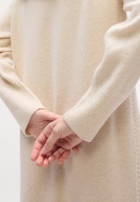 Personne portant un long pull crème avec les manches couvrant les poignets, mains délicatement jointes derrière le dos.
