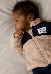 Fleece-gevoerd jack, beige en marineblauw, met een ritssluiting en een "LET'S GO" patch. Glad textuur met een gezellige pasvorm, geschikt voor zuigelingen.
