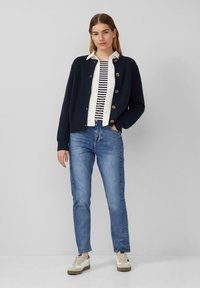 Marineblauer gerippter Cardigan mit Holzknöpfen, getragen über einem weiß-schwarz gestreiften Shirt. Kombiniert mit hellblauen Jeans und tanfarbenen Sneakers.
