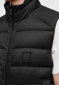 Мъж, облечен с черен квадратиран безръкавен елек Belstaff с цип върху бяла риза с къс ръкав, показващ детайли на гърдите и врата.