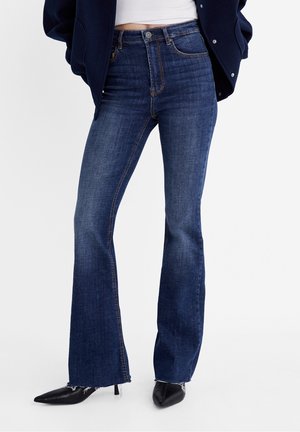 Donkerblauwe flared jeans met een hoge taille, voorzien van contrasterende stiksels, zakken en rafelige zomen, gecombineerd met puntige zwarte hakken.