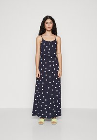 Robe maxi marine à pois blancs, bretelles spaghetti, taille cintrée et jupe fluide. Associée à des sandales vert lime.