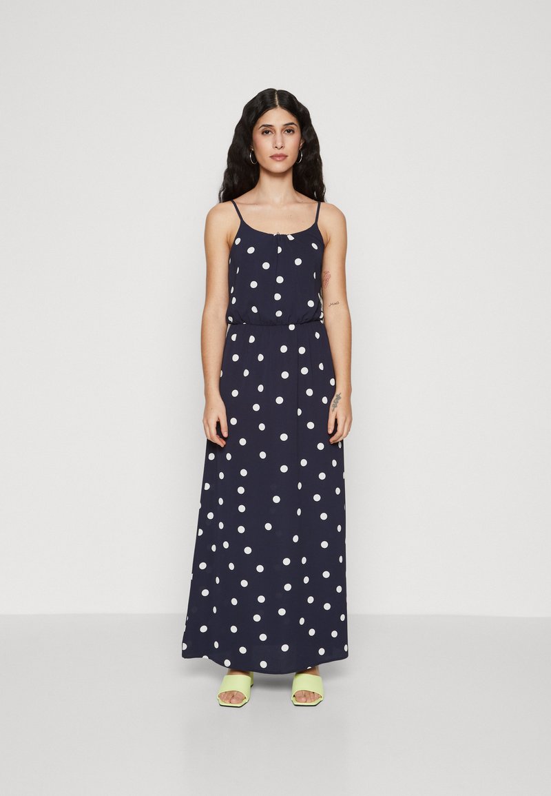 Robe maxi marine à pois blancs, bretelles spaghetti, taille cintrée et jupe fluide. Associée à des sandales vert lime.