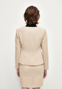Blazer beige su misura con taglio strutturato, tessuto testurizzato e colletto nero, abbinato a una gonna attillata coordinata con spacco posteriore.