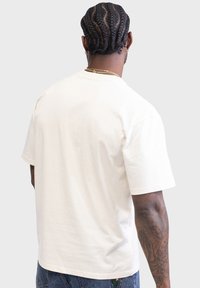 Cream-colored cotton t-shirt, short sleeves, crew neck, relaxed fit, smooth texture. Visible tattoos on the arm.