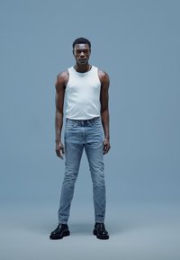 Balts bezroku tops, gaiši zilas džinsu bikses un melni potīšu zābaki. Vienkāršs dizains bez redzamām rakstām. Neitrāla fona.