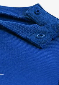 Guler din material albastru cu țesătură texturată, prevăzut cu două închizători cu capse albastre din plastic și suprafață netedă. Este vizibil un detaliu de cusături albe.