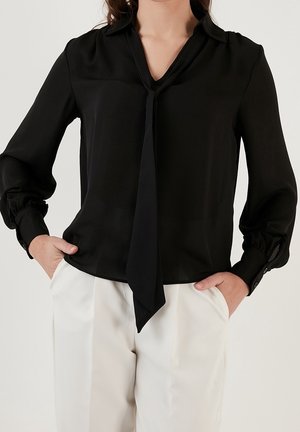 Kvinde iført en sort bluse med lange ærmer og bindebånd foran i halsen, kombineret med hvide bukser med høj talje, hænderne puttet i lommerne.