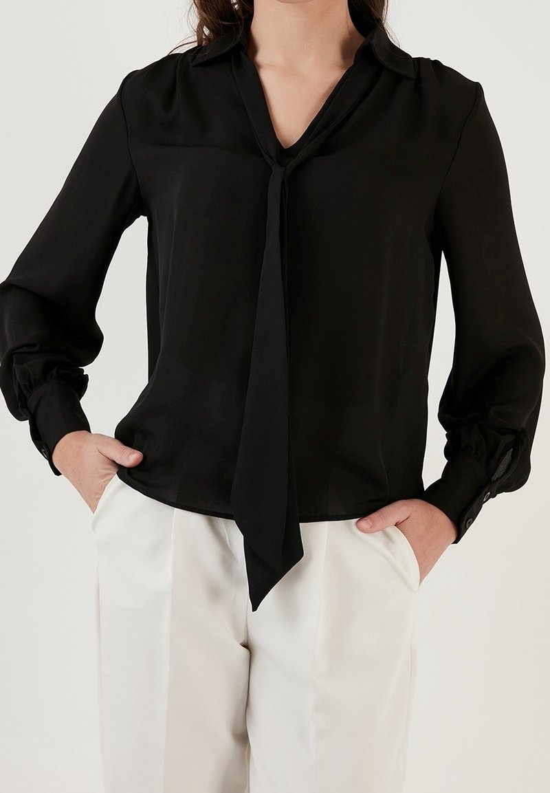 Femme portant un chemisier noir à manches longues avec un nœud à l'avant du col, associé à un pantalon blanc taille haute, mains dans les poches.