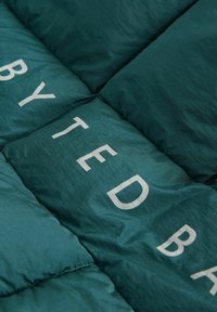 Zeleno prešito blago z gladko teksturo, na katerem so velike bele črke, ki tvorijo besedo "BYTED". Sodobna in elegantna zasnova.